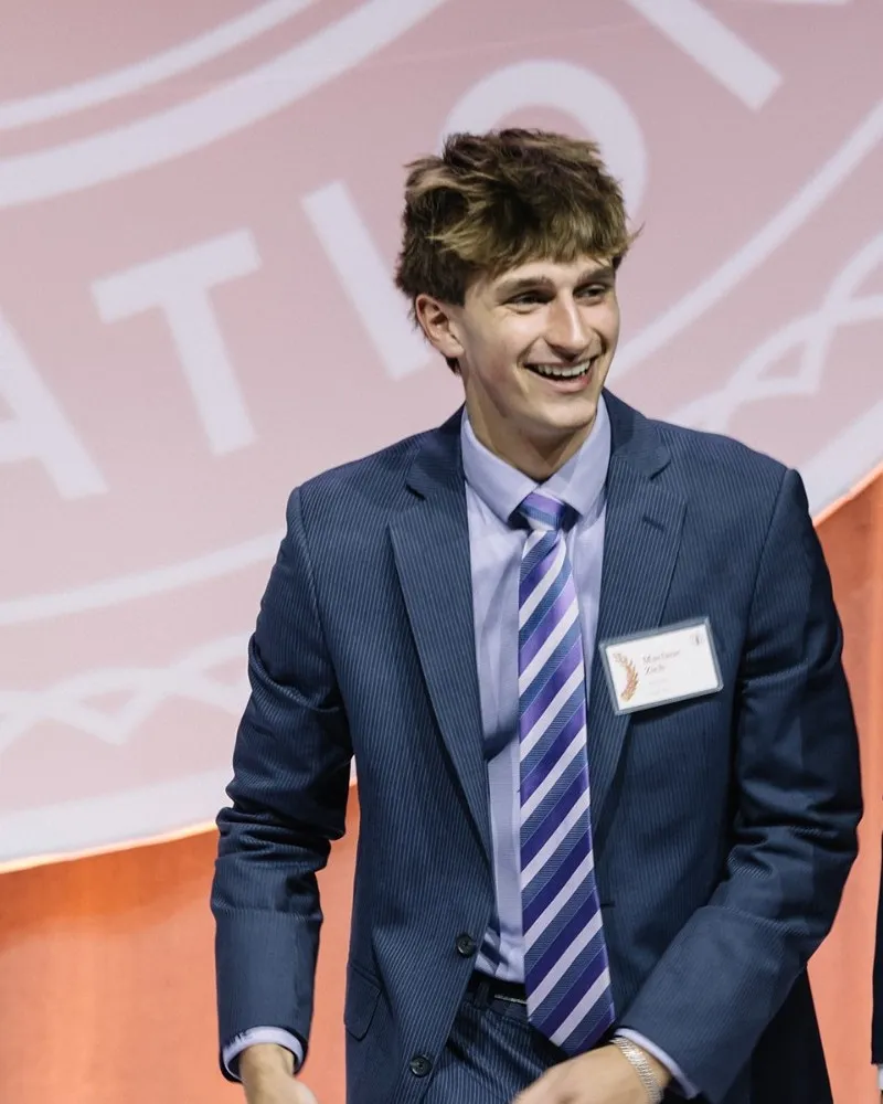 Maclane Zich smiling on stage in a navy suit at the Coca-Cola Scholars Foundation event