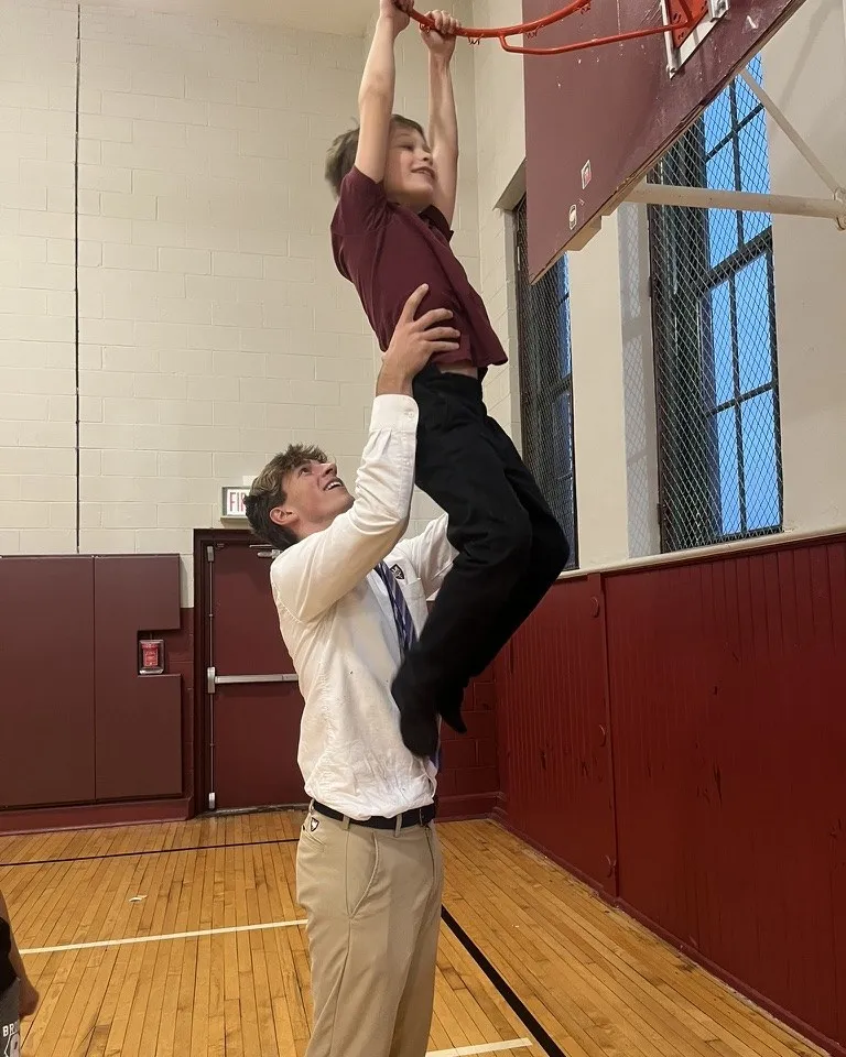 Maclane Zich lifting a student up to grab a basketball rim in a school gym